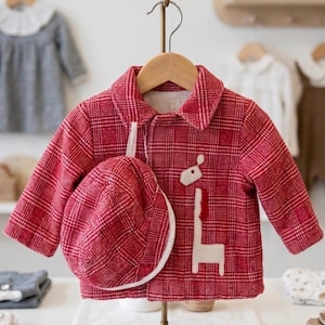 May include: A red plaid jacket and matching cap for a child. The jacket features a giraffe applique and is displayed on a hanger. The cap has a white brim. Other children's clothing is visible in the background.