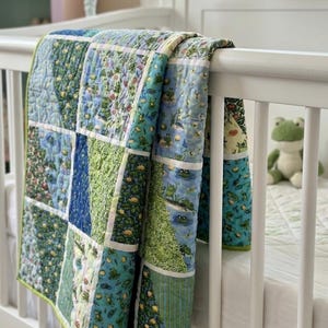 May include: A patchwork baby quilt with a frog theme. The quilt features squares of blue and green fabrics with frog and lily pad patterns. The quilt is draped over a white crib rail, with a stuffed frog toy visible.