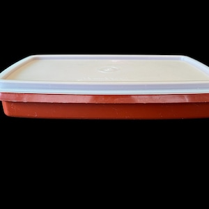 May include: A rectangular food storage container with a white lid and a red base. The lid has a white rim and a slightly raised center. The container is made of plastic and is likely used for storing food.