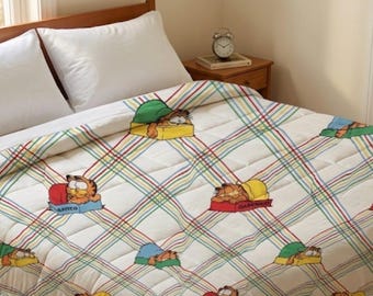Puede incluir: Una colcha blanca acolchada con un patrón repetido de Garfield el gato durmiendo en camas de varios colores. La colcha tiene un diseño a cuadros con líneas rojas, verdes y amarillas.