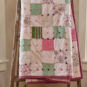 May include: A handmade patchwork quilt with a variety of floral, polka dot, and solid fabric squares in white, green, pink, and purple. The quilt is draped over a wooden ladder. The quilt has a pink border and is decorated with small, colorful pom-poms.