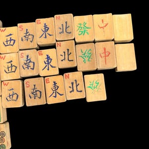 May include: A collection of rectangular wooden mahjong tiles arranged in rows. The tiles feature engraved Chinese characters painted in blue, green, and red, along with small English directional letters W, S, E, and N printed above the characters.
