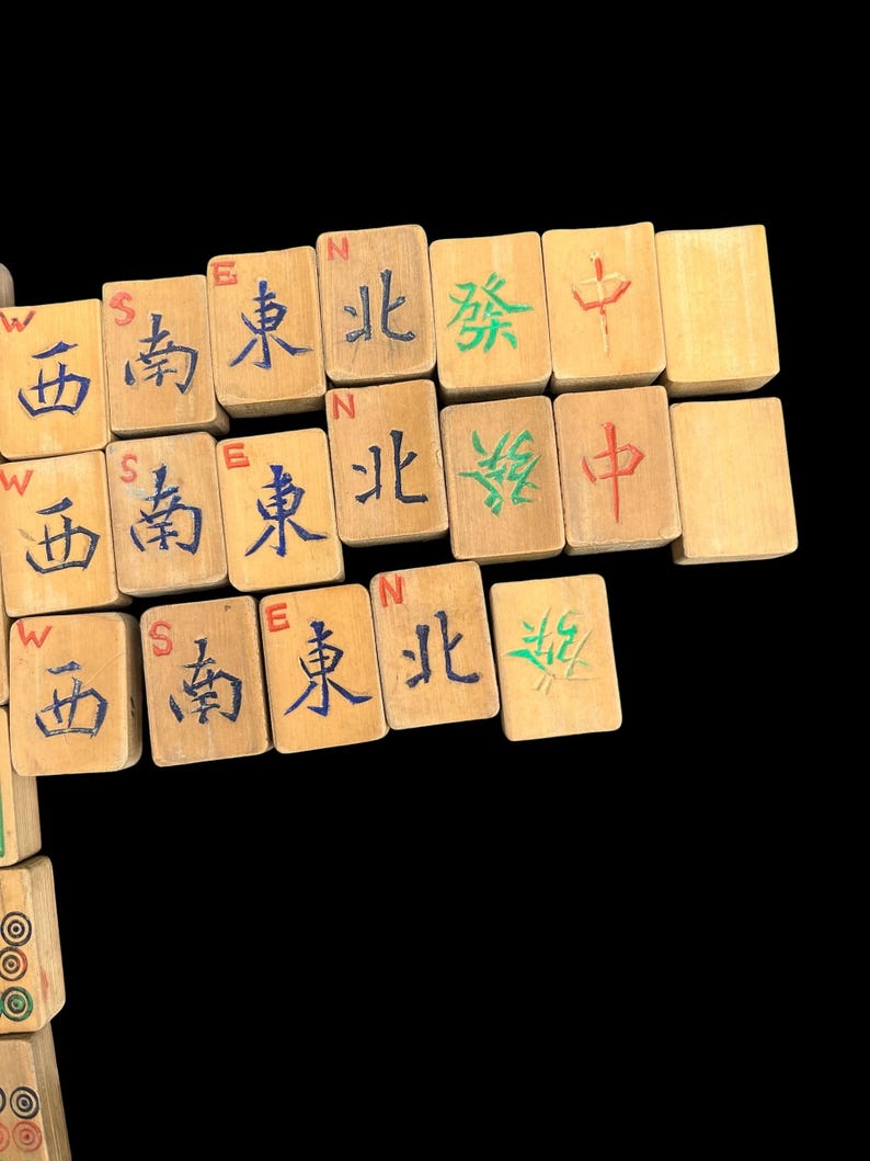 May include: A collection of rectangular wooden mahjong tiles arranged in rows. The tiles feature engraved Chinese characters painted in blue, green, and red, along with small English directional letters W, S, E, and N printed above the characters.