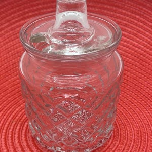 May include: A clear glass jar with a lid and a decorative diamond pattern on the body. The jar has a small handle on the lid and is sitting on a red surface. The jar is designed to hold small items.