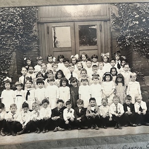 May include: A black and white vintage photograph of a large group of children posed in front of a building entrance with ivy-covered walls. The children are arranged in rows, wearing formal attire including dresses, collared shirts, and ties.