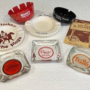 May include: Collection of vintage ashtrays featuring various business names and logos. The ashtrays are made of glass and ceramic and feature a variety of colors and designs. The names include "Harry Starker's on the Plaza", "Mc Putsche's Prime Dining", "The Smoke Stack BBQ", "Spaghetti Spot", "Waid's Family Restaurant", and "The Golden Ox Restaurant & Cocktail Lounge".