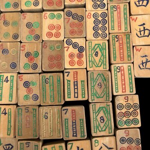 May include: A collection of rectangular wooden Mahjong tiles arranged in a grid. Each tile features hand-painted designs in red, green, blue, and black, including numeric characters, bamboo patterns, and circular motifs, with some tiles showing the letter W.