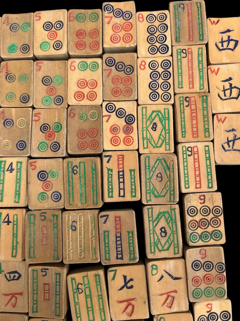 May include: A collection of rectangular wooden Mahjong tiles arranged in a grid. Each tile features hand-painted designs in red, green, blue, and black, including numeric characters, bamboo patterns, and circular motifs, with some tiles showing the letter W.