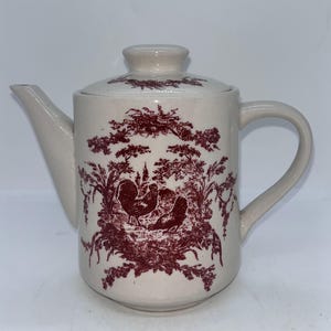 May include: A white ceramic teapot with a red floral and bird design. The teapot has a lid and a handle.