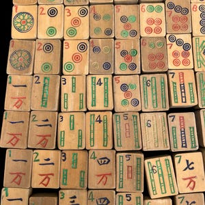 May include: A collection of rectangular wooden mahjong tiles arranged in a grid. Each tile features hand-painted numerals, circular patterns, and bamboo symbols in red, green, blue, and black ink on a light tan wood surface with visible grain.