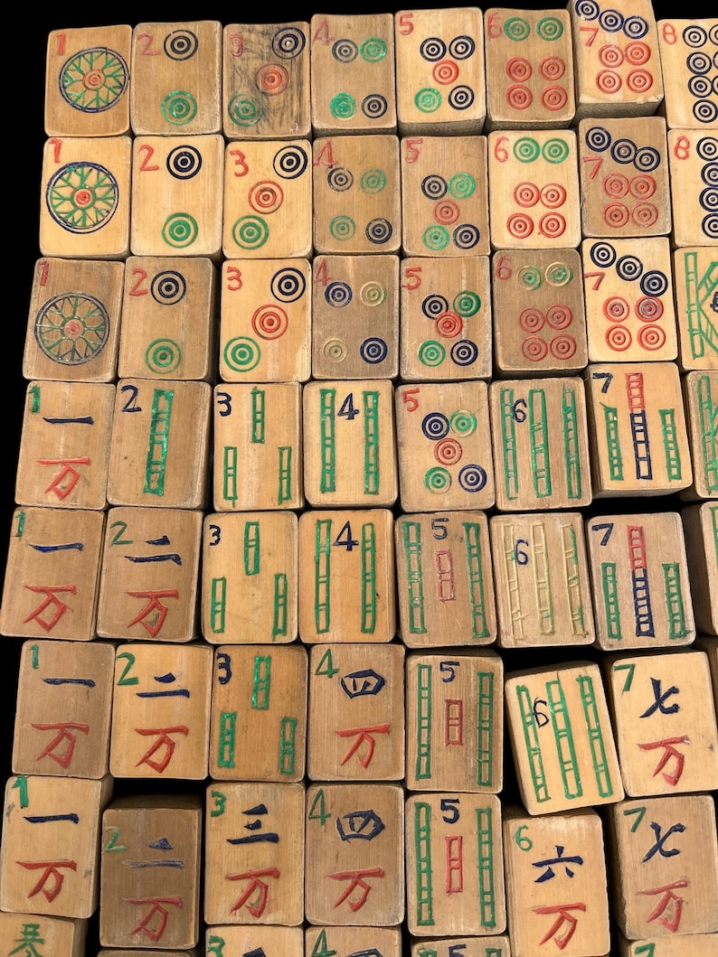 May include: A collection of rectangular wooden mahjong tiles arranged in a grid. Each tile features hand-painted numerals, circular patterns, and bamboo symbols in red, green, blue, and black ink on a light tan wood surface with visible grain.