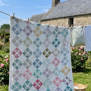 May include: A white quilt with colorful diamond-shaped patches hanging on a clothesline. The quilt features various floral patterns in shades of pink, blue, green, and yellow. The background includes a stone building and a clear blue sky.