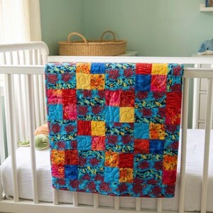 May include: A colorful patchwork quilt draped over a white crib. The quilt features squares of red, blue, yellow, and orange fabrics with various patterns. The crib is in a room with a light green wall and a window.