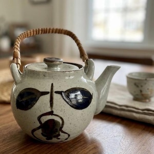 May include: A ceramic teapot with a woven handle and a matching teacup. The teapot is off-white with dark blue and black painted details. The teacup is small and off-white. The teapot and teacup are on a wooden table.