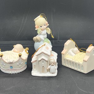May include: A collection of Precious Moments figurines. The figurines include a baby in a crown-shaped cradle, an angel sitting on a globe, a church with an angel, and a baby in a crib. Each figurine has a gold string for hanging.