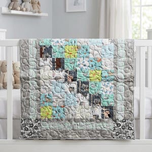 May include: A baby quilt with a patchwork design in shades of gray, blue, and yellow. The quilt features a variety of animal-themed squares and a decorative border. The quilt is displayed on a white crib.