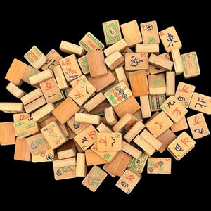 May include: A collection of rectangular wooden Mahjong tiles scattered in a pile. Each tile features a light brown wood grain finish with various engraved symbols, Chinese characters, and numbers painted in red, green, blue, and black ink colours.