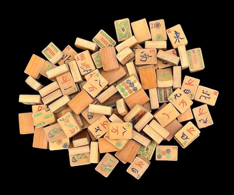 May include: A collection of rectangular wooden Mahjong tiles scattered in a pile. Each tile features a light brown wood grain finish with various engraved symbols, Chinese characters, and numbers painted in red, green, blue, and black ink colours.