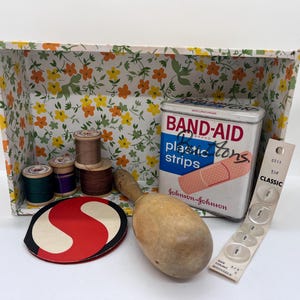 May include: A vintage floral patterned box filled with sewing supplies, including spools of thread, a wooden thimble, a red and white fabric circle, and a tin of Band-Aid plastic strips.