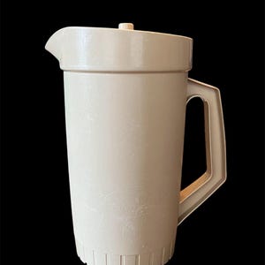 May include: A beige plastic pitcher with a lid and handle. The pitcher has a textured surface and a spout for pouring. The handle is rectangular and attached to the side of the pitcher. The lid has a small knob on top.