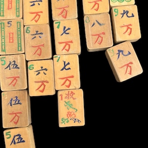 May include: A collection of rectangular wooden Mahjong tiles featuring hand-painted Chinese characters, numbers, and symbols in red, blue, green, and black ink. The tiles display various numerical values and suit markings, including bamboo patterns and circular designs.