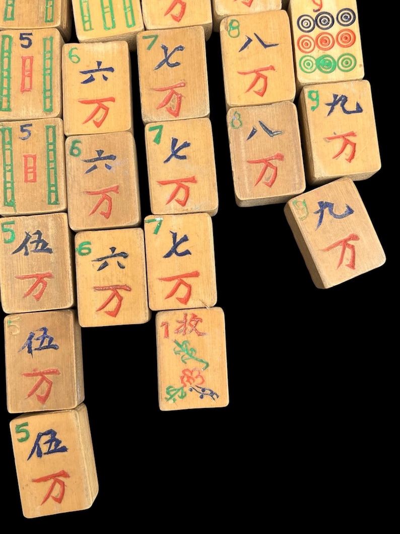 May include: A collection of rectangular wooden Mahjong tiles featuring hand-painted Chinese characters, numbers, and symbols in red, blue, green, and black ink. The tiles display various numerical values and suit markings, including bamboo patterns and circular designs.