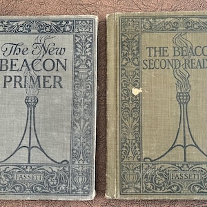 May include: Two vintage books, "The New Beacon Primer" and "The Beacon Second Reader," with ornate covers. The covers feature a stylized torch design and decorative borders. The books are in shades of gray and tan, with the publisher's name, "Fassett," at the bottom.