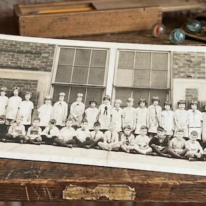 May include: A vintage black and white panoramic photograph depicting a large group of children and an adult standing and sitting in front of a brick school building with large windows. The photograph is rectangular with a white border.