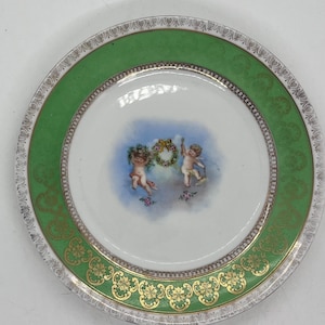May include: A decorative porcelain plate with a green and gold border. The plate features a central image of two cherubs with a floral wreath. The outer rim has a gold and white floral design.