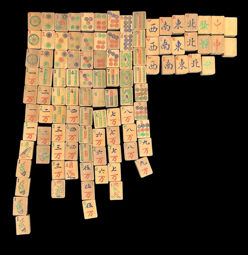May include: A collection of rectangular light-coloured wooden Mahjong tiles arranged in a grid pattern. Each tile features hand-painted Chinese characters, numbers, and symbols in red, green, blue, and black ink, representing various suits and honours.