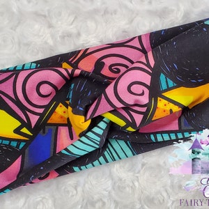 Op de afbeelding: Een kleurrijke haarband met een glas-in-lood-achtig ontwerp. Het patroon bevat roze, gele, blauwe en turquoise vormen op een zwarte achtergrond. De haarband is aan de bovenkant geknoopt en is waarschijnlijk gemaakt van zachte stof.