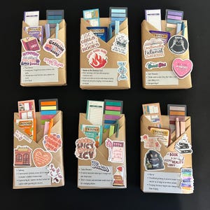 May include: Six brown paper packages wrapped with colorful stickers and book-themed decorations. Each package includes a book-themed sticker, a tea bag, and a set of colorful sticky notes. The stickers include phrases like "Hot Girls Read Books", "Emotionally Fictional Characters", "A Book A Day Keeps Reality Away", and "Caffeine Smutty Books".