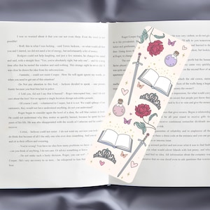 May include: A bookmark with a white background and a variety of illustrations, including a sword, a crown, a book, a potion bottle, a rose, and butterflies. The bookmark has a gold glitter effect.