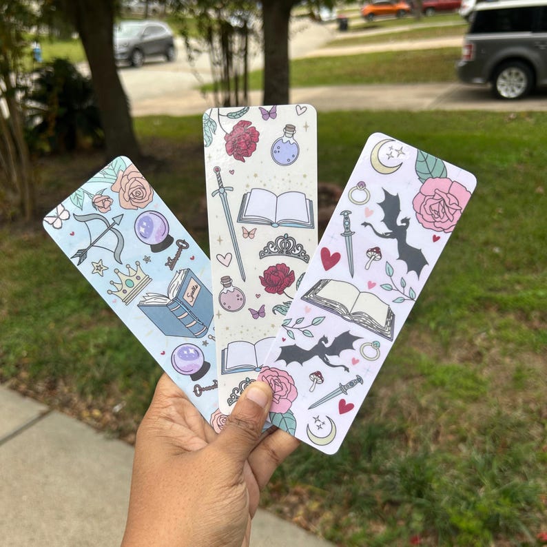 May include: Three colorful bookmarks with whimsical illustrations. The bookmarks feature a dragon, a book, a crown, a key, a potion bottle, a sword, a butterfly, a crystal ball, and a ring. The bookmarks are printed on white cardstock.