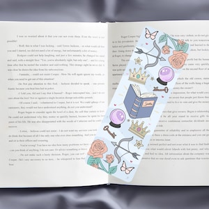 May include: A blue bookmark with a variety of illustrations, including a book, a crown, a key, a butterfly, an arrow, and a crystal ball. The bookmark has the word "Spells" written on the book. The bookmark is placed on top of an open book with text.