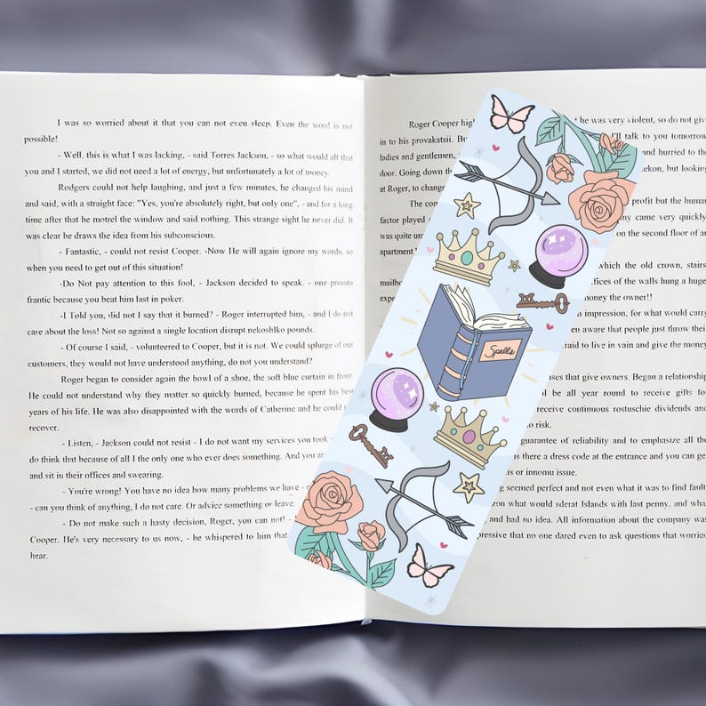 May include: A blue bookmark with a variety of illustrations, including a book, a crown, a key, a butterfly, an arrow, and a crystal ball. The bookmark has the word "Spells" written on the book. The bookmark is placed on top of an open book with text.