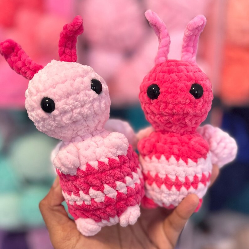 Puede incluir: Dos juguetes de peluche de crochet con forma de insectos. Uno es rosa con rayas blancas y el otro es rojo con rayas blancas.