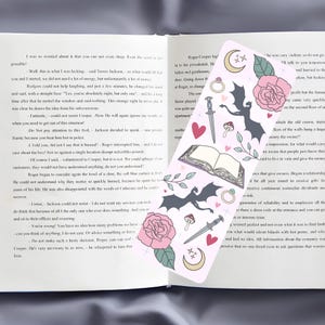 May include: A pink bookmark with a black dragon, a book, a dagger, a ring, a mushroom, a heart, a moon, and stars. The bookmark has the text "Roger Cooper High" and "The apartment was quite un" on it.
