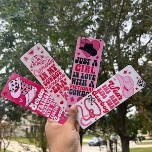 May include: Four pink and white bookmarks with cowboy-themed designs. Each bookmark features text such as "Currently in my cowboy romance era" and "Just a girl in love with a fictional cowboy." The bookmarks have cowboy hat and heart illustrations.