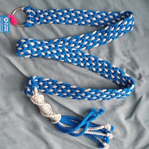 Puede incluir: Un cinturón largo trenzado con un patrón repetitivo de cordón azul real y gris plateado. El cinturón está asegurado en un extremo con un gran anillo de metal plateado y terminado en el otro extremo con un nudo retorcido y una borla.