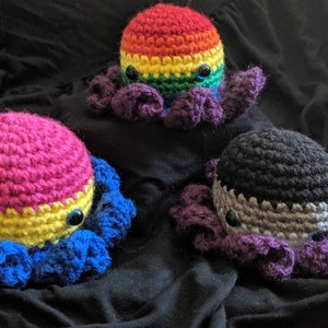 May include: Three crocheted octopus plushies with different coloured bodies and tentacles. The octopus on the left has a pink, yellow, and blue body. The octopus in the middle has a rainbow coloured body. The octopus on the right has a black, grey, and purple body.