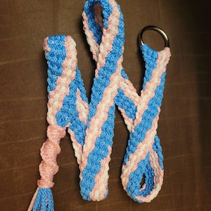 Op de afbeelding: Een blauw en roze gevlochten koord met een zilveren ring en een kwast. Het koord is gemaakt van twee kleuren koord, die in een diagonaal patroon met elkaar zijn verweven.