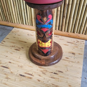 May include: A wooden tiki bar stool with a red cushioned seat. The stool is carved with a tiki face and painted with blue, red, and yellow accents. The stool is sitting on a bamboo mat.