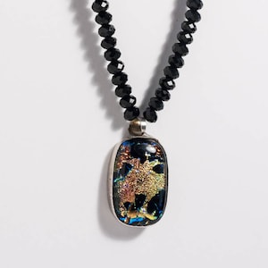 May include: A black beaded necklace with a rectangular pendant. The pendant has a silver frame and features a colorful, abstract design with gold, blue, and red hues. The necklace is displayed against a white background.