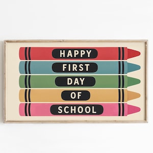 May include: A framed print featuring five colorful crayons with the words "HAPPY FIRST DAY OF SCHOOL" in white text on black ovals. The crayons are red, teal, green, yellow, and pink, set against a cream background.