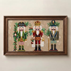 Op de afbeelding: Een rechthoekig ingelijst schilderij met drie naast elkaar staande notenkrakerfiguren. De linkerfiguur draagt groen, de middelste figuur rood met een gouden kroon en de rechterfiguur blauw, allemaal versierd met groen.