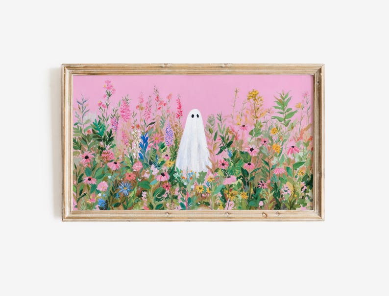 May include: A framed art print featuring a white ghost figure standing in a field of colorful wildflowers. The artwork has a pink background and is set in a light brown wooden frame. The flowers are in shades of pink, purple, yellow, and green.