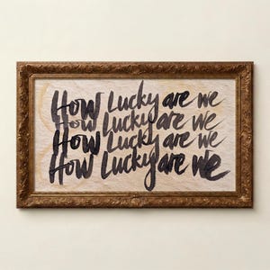 May include: A framed artwork with the phrase "How Lucky are we" written in black calligraphy. The artwork has a light-colored background and is encased in a decorative, ornate, gold-toned frame. The artwork is displayed against a neutral wall.