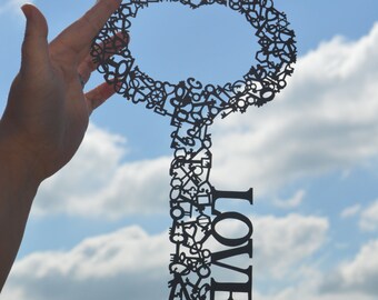 May include: A black silhouette of a key made of many small keys, with the word "LOVE" written on the bottom of the key. The key is held up against a blue sky with white clouds.