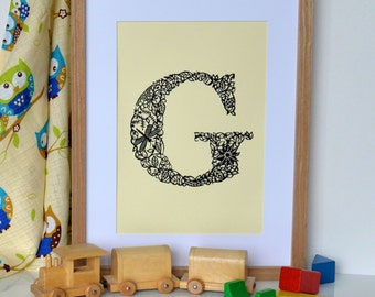 May include: A framed print of the letter G, made from black paper cut-outs of leaves and flowers, on a pale yellow background. The frame is made of light wood and is sitting on a white surface with a toy train and wooden blocks.